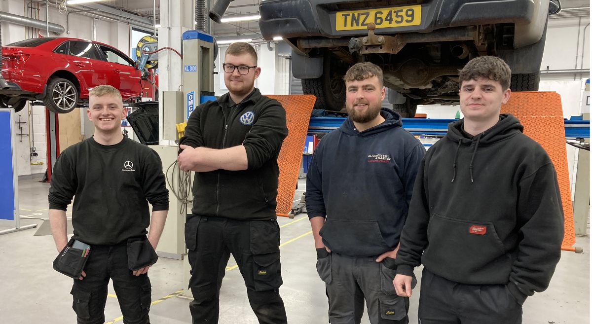 Four male apprentices in work wear smiling to camera with elevated vehicles in garage in background.